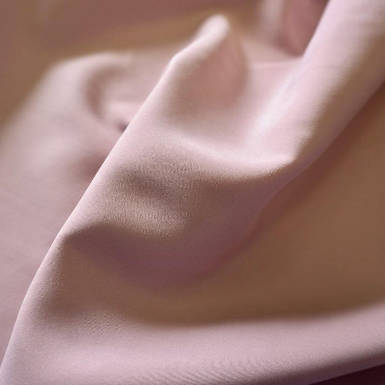 May include: Close-up of a soft, flowing fabric in a muted rose color. The material is draped, creating gentle folds and shadows that highlight its texture and sheen. The fabric appears smooth and luxurious.