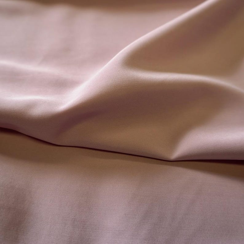 May include: Close-up of a soft, flowing fabric in a muted rose color. The fabric has subtle folds and shadows, creating a sense of depth and texture. The material appears smooth and luxurious, suggesting a high-quality textile.