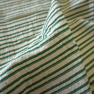May include: A close-up shot of a white fabric with green and black horizontal stripes. The fabric is textured and folded, creating shadows. The stripes are evenly spaced. The overall effect is of a textured textile.