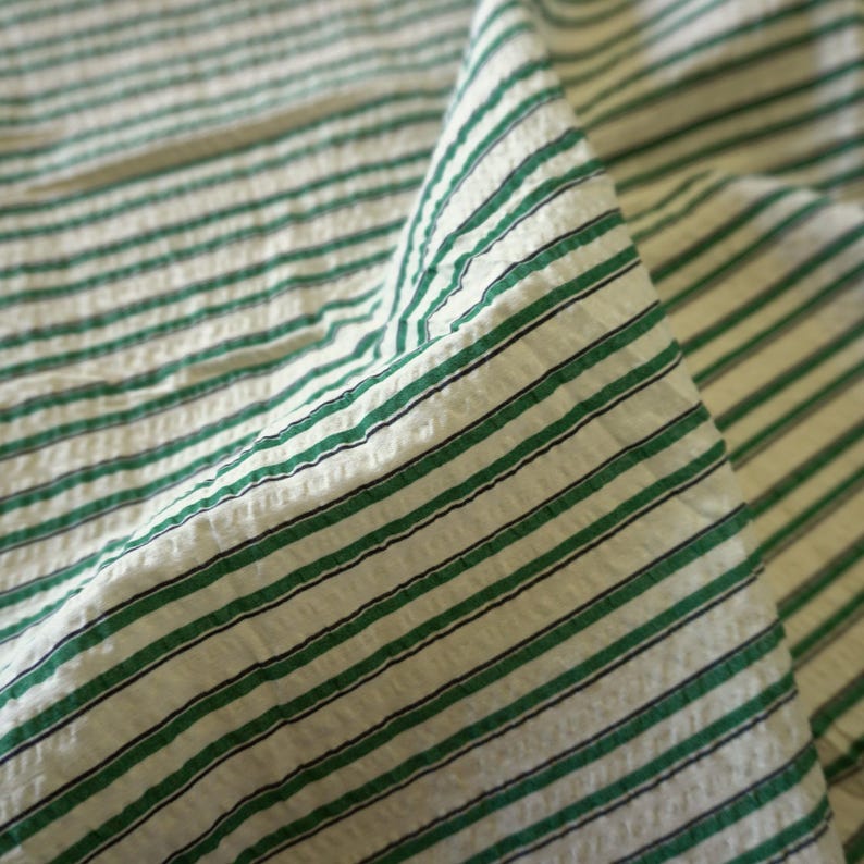 May include: A close-up shot of a white fabric with green and black horizontal stripes. The fabric is textured and folded, creating shadows. The stripes are evenly spaced. The overall effect is of a textured textile.