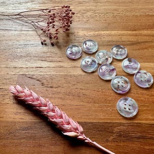 May include: Iridescent buttons with a swirl of purple and white are scattered on a wooden surface. A pink wheat stalk and dried floral elements are also present. These buttons are suitable for sewing and craft projects.