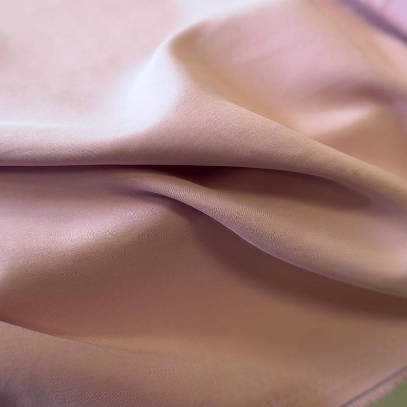 May include: Close-up of a soft, flowing fabric in a muted rose color. The material has a subtle sheen, with gentle folds and shadows creating depth. The fabric appears smooth and luxurious, suggesting a high-quality textile.