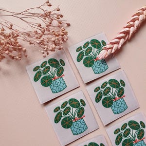 Pode incluir: Seis patches bordados para passar a ferro que representam uma planta verde com folhas redondas num vaso azul com uma fita rosa. Os patches est&atilde;o sobre um fundo rosa claro.