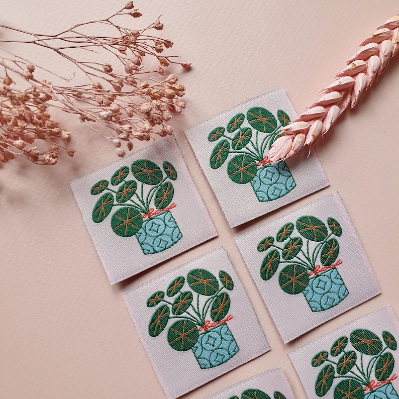 Pode incluir: Seis patches bordados para passar a ferro que representam uma planta verde com folhas redondas num vaso azul com uma fita rosa. Os patches est&atilde;o sobre um fundo rosa claro.