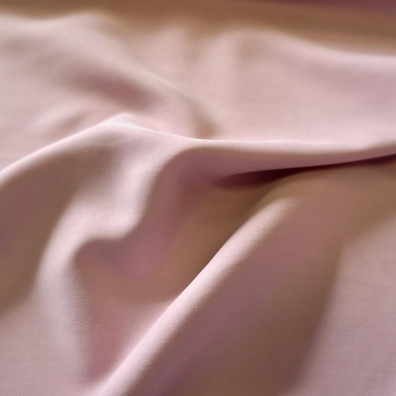 May include: Close-up of a soft, flowing fabric in a light mauve color. The material has gentle folds and shadows, creating a sense of depth and texture. The fabric appears smooth and possibly silky, ideal for apparel or home decor.