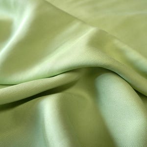 May include: Close-up of a light green fabric with a ribbed texture. The fabric is folded, creating shadows and highlights that emphasize its texture and drape. The color is a soft, muted green.
