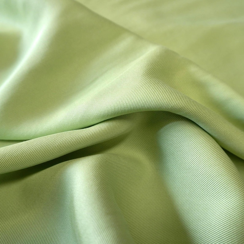 May include: Close-up of a light green fabric with a ribbed texture. The fabric is folded, creating shadows and highlights that emphasize its texture and drape. The color is a soft, muted green.