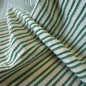 May include: Close-up of a folded fabric featuring a white base with green and black stripes. The fabric has a textured surface, with the stripes running diagonally. The material is likely cotton or linen, suitable for clothing or home textiles.
