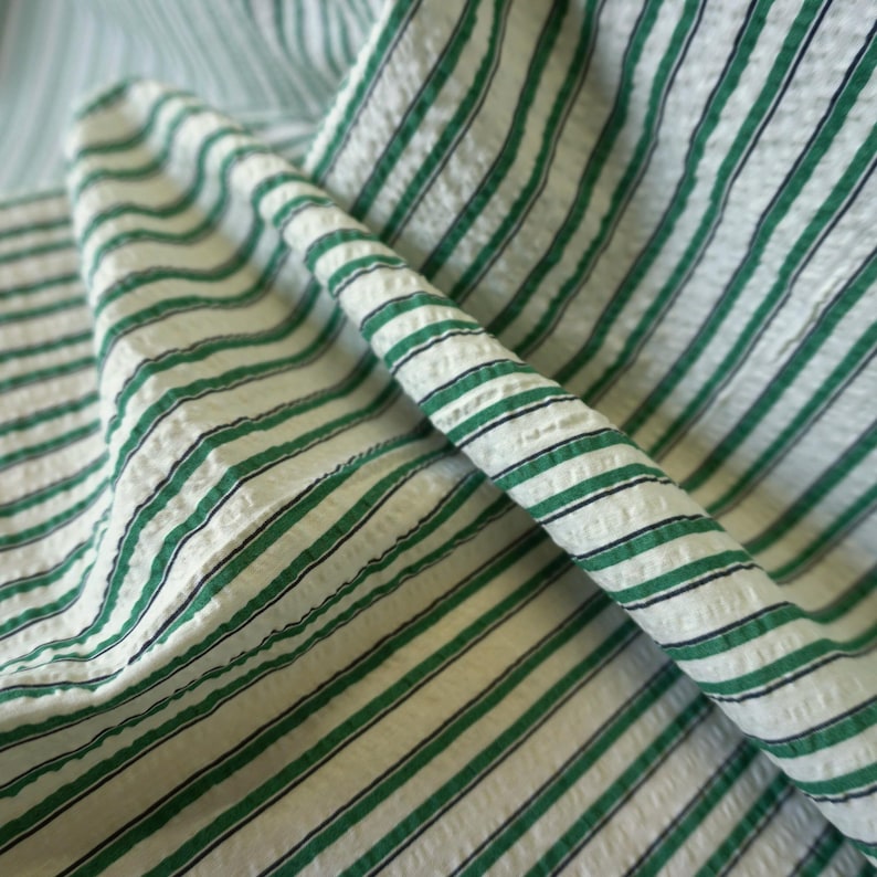 May include: Close-up of a folded fabric featuring a white base with green and black stripes. The fabric has a textured surface, with the stripes running diagonally. The material is likely cotton or linen, suitable for clothing or home textiles.