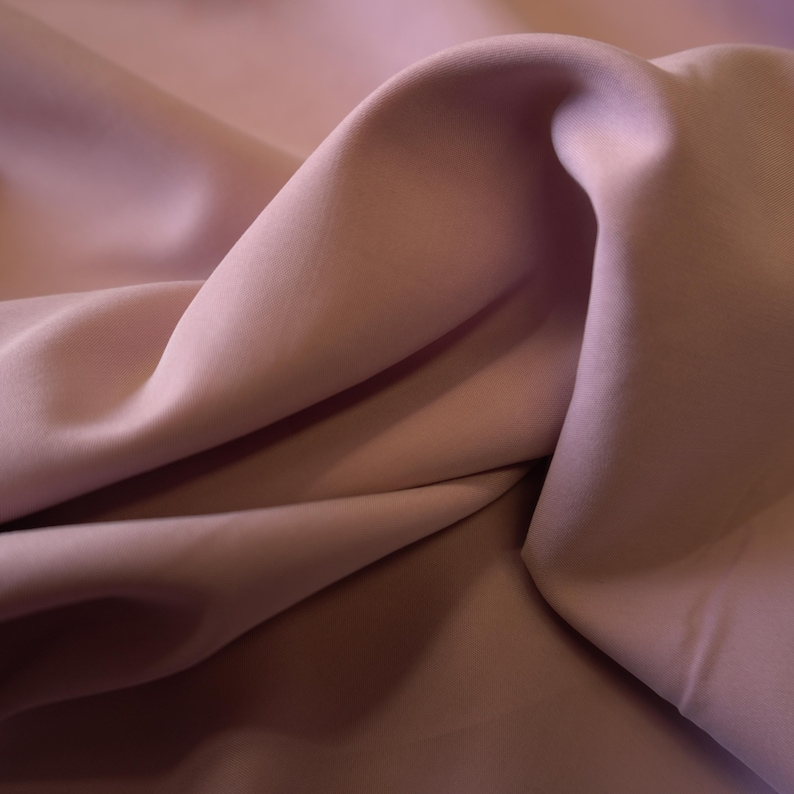 May include: Close-up of a flowing fabric in shades of mauve and light brown. The material is soft and draped, with visible folds and shadows that create depth and texture. The fabric appears to be smooth and luxurious.