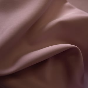 May include: Close-up of a flowing fabric in a muted mauve color. The material is smooth and appears to be draped, with soft shadows and highlights creating depth. The fabric's texture is fine and delicate, suggesting a luxurious textile.