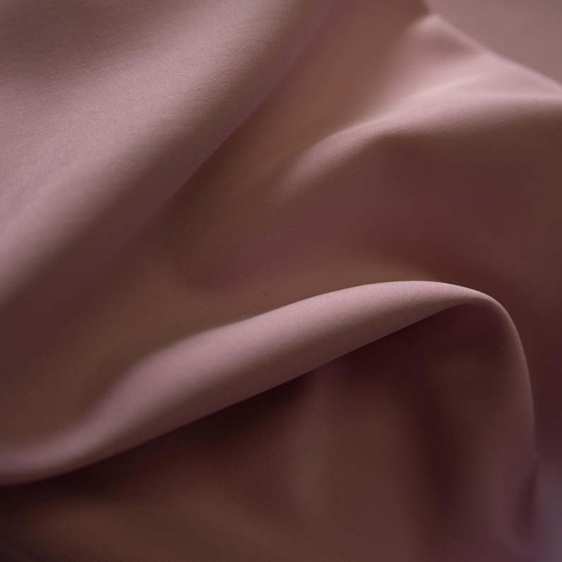 May include: Close-up of a flowing fabric in a muted mauve color. The material is smooth and appears to be draped, with soft shadows and highlights creating depth. The fabric's texture is fine and delicate, suggesting a luxurious textile.