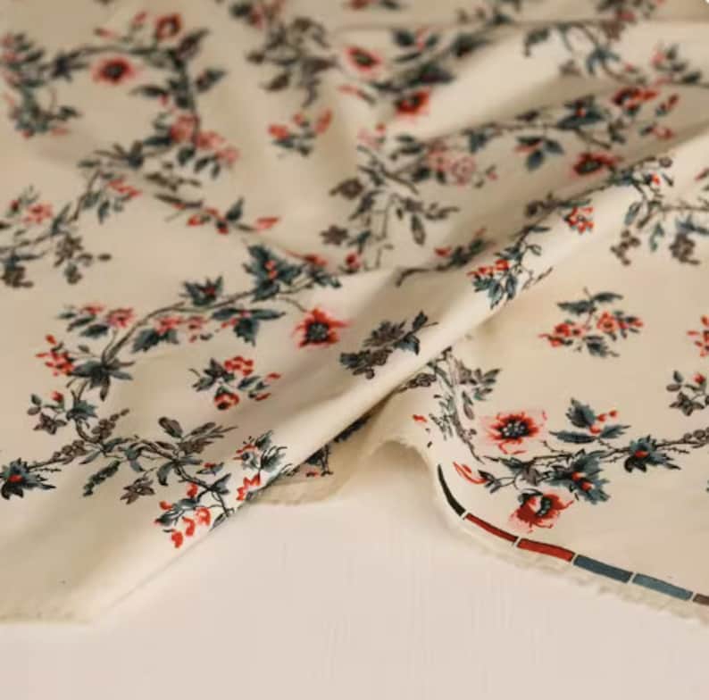May include: Cream-colored fabric with a repeating floral pattern. The flowers are red, pink, and blue with green leaves. The fabric is folded in half and the edge is trimmed with a red and blue stripe.