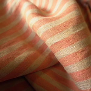 May include: Close-up of a fabric with a repeating pattern of peach and off-white stripes. The fabric is folded, showing the texture and direction of the stripes. The material appears to be linen or a similar woven textile.