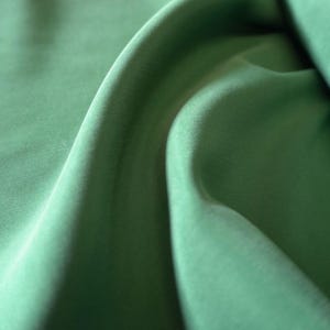 May include: Close-up of a flowing, sage green fabric. The material has a soft sheen and is draped in gentle folds, creating shadows and highlights. The fabric appears smooth and luxurious, suggesting a high-quality textile.