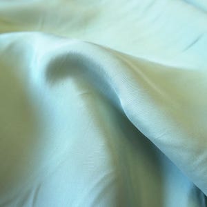 May include: Close-up of a light blue fabric with soft folds and subtle texture. The fabric has a smooth, slightly shiny appearance, with variations in tone creating depth and dimension. The overall effect is one of gentle movement and fluidity.