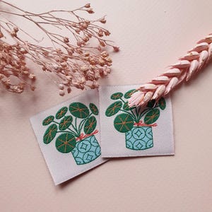 Pode incluir: Dois patches para passar a ferro com uma planta verde em vaso com um vaso de padr&atilde;o azul e branco. A planta tem folhas redondas, verdes com acentos laranja. Os patches est&atilde;o sobre um fundo rosa.