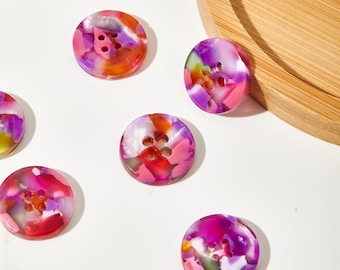 Puede incluir: Una colección de botones redondos y coloridos con cuatro agujeros. Los botones presentan un diseño veteado en tonos rosa, morado, naranja y blanco. Están dispersos sobre una superficie blanca, con una bandeja de madera y detalles florales en el fondo.