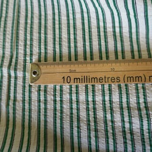 May include: Close-up of a textured fabric with vertical stripes in white, green, and dark blue. A wooden ruler with millimeter markings is placed on the fabric.