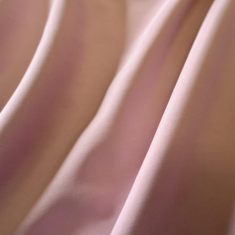 May include: Close-up of soft, flowing fabric in shades of light pink and peach. The material is draped, creating gentle folds and shadows that highlight its texture and sheen. The overall effect is smooth and elegant.