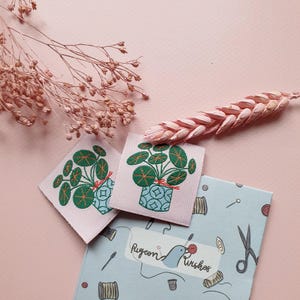 Pode incluir: Dois remendos de tecido rosa com um desenho bordado de uma planta verde em um vaso azul. Os remendos est&atilde;o sobre um fundo azul com um p&aacute;ssaro branco e o texto "Pigeon Wishes".