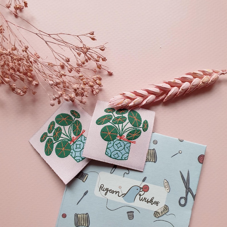 Pode incluir: Dois remendos de tecido rosa com um desenho bordado de uma planta verde em um vaso azul. Os remendos est&atilde;o sobre um fundo azul com um p&aacute;ssaro branco e o texto "Pigeon Wishes".