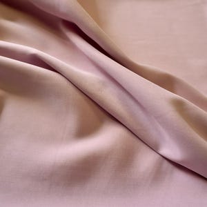 May include: Close-up of a soft, flowing fabric in a light pink hue. The material is draped, creating folds and shadows that highlight its texture and drape. The fabric appears smooth and likely suitable for clothing or home decor.