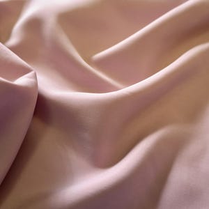 May include: Close-up of a flowing, light pink fabric. The material is smooth and appears to be silk or a similar luxurious textile. The folds and shadows create a sense of depth and texture, highlighting the fabric's drape and sheen.