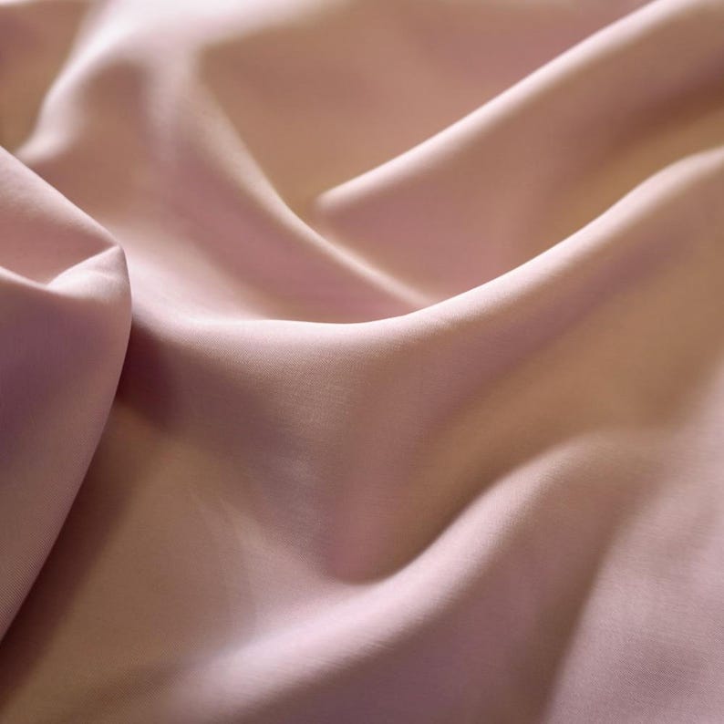 May include: Close-up of a flowing, light pink fabric. The material is smooth and appears to be silk or a similar luxurious textile. The folds and shadows create a sense of depth and texture, highlighting the fabric's drape and sheen.