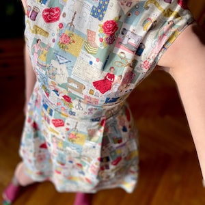 Puede incluir: Vestido sin mangas con un diseño de patchwork con ilustraciones de costura. El vestido tiene un escote redondo y una falda cruzada. La tela es una mezcla de azul claro, blanco y rosa, con imágenes de máquinas de coser, patrones y rosas.