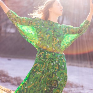 May include: A woman wearing a green, patterned dress with a long, flowing skirt and a cinched waist. The dress has a sheer, almost see-through fabric and is adorned with a belt.
