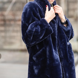 Op de afbeelding: Een lange marineblauwe imitatiebontjas met een knoopsluiting en twee zakken. De jas heeft brede revers en lange mouwen. Het materiaal lijkt zacht en de jas heeft een diepe blauwe tint.