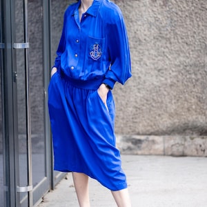 Puede incluir: Un vestido vintage azul vibrante, compuesto por una blusa de manga larga y una falda a juego. La blusa presenta un pequeño emblema bordado y botones dorados. El conjunto se completa con tacones verdes oscuros. El vestido es de estilo a la rodilla.