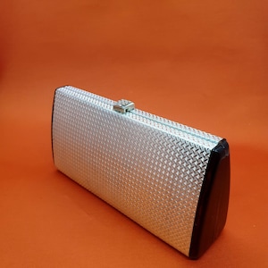 Puede incluir: Un bolso de mano rectangular con un exterior plateado y un acabado texturizado de patrón de diamantes. El bolso presenta un panel lateral negro sólido y un pequeño cierre metálico situado en el borde superior del marco para asegurar.