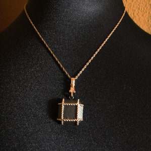 Könnte beinhalten: Eine goldene Kettenkette mit einem schwarzen quadratischen Anhänger. Der Anhänger ist von einem goldenen Drahtrahmen umgeben.
