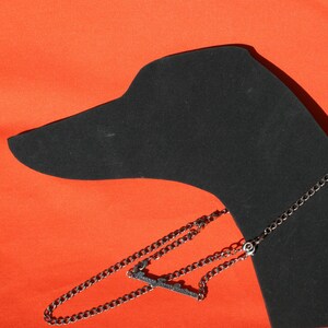 Könnte beinhalten: Eine schwarze Silhouette eines Hundekopfes mit einer silbernen Kettenkette und einem dekorativen Anhänger. Der Hintergrund ist einfarbig orange.