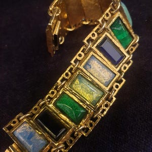 Könnte beinhalten: Goldfarbenes Armband mit rechteckigen, farbigen Emaille-Einsätzen. Das Armband hat ein geometrisches Design mit grüner, blauer und schwarzer Emaille. Die Glieder sind durch kleine Ketten verbunden. Das Armband wird auf einer dunklen Oberfläche präsentiert.
