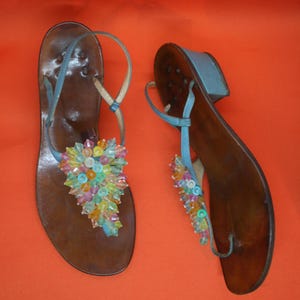 May include: A pair of brown leather sandals with blue straps and colorful beaded embellishments. The sandals have a thong style design with a small heel. The beaded accents feature a variety of colors, including pink, yellow, and blue.