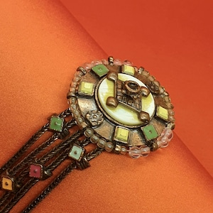 Könnte beinhalten: Ein Armband im Vintage-Stil mit einem kreisförmigen, verzierten Mittelstück. Das Mittelstück hat ein cremefarbenes Emaille-Design mit grünen und gelben quadratischen Akzenten. Das Armband hat mehrere Stränge aus Kettengliedern und farbige quadratische Charms.