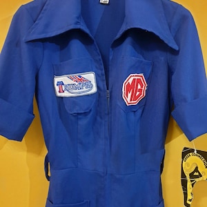 Könnte beinhalten: Ein blauer Vintage-Overall mit kurzen Ärmeln, einem Kragen und einem Reißverschluss vorne. Er hat zwei Vordertaschen und Aufnäher mit den Logos Triumph und MG. Der Overall hat eine taillierte Silhouette und einen Gürtel.