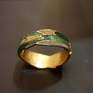 Puede incluir: Brazalete dorado con bisagras y parte superior decorativa. La parte superior presenta un diseño ondulado con detalles verdes, dorados y plateados. El brazalete tiene una superficie texturizada y un cierre de broche.