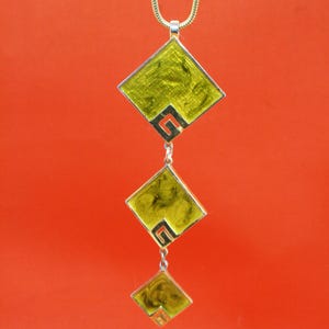Puede incluir: Un collar con colgante que presenta tres colgantes cuadrados de color verde lima con bordes plateados y una cadena plateada. Los colgantes están conectados por pequeños eslabones plateados y cuelgan sobre un fondo naranja liso. El collar tiene un diseño geométrico.