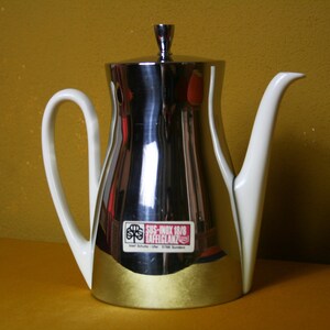 May include: A silver and white stainless steel coffee pot with a white handle and spout. The pot has a label that reads "SUS-INOX 18/8 TAFELGLANZ".