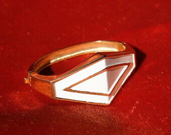 May include: A gold bangle bracelet with a white enamel geometric design. The design features a white triangle with a gold outline.