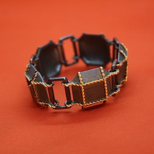 Puede incluir: Un brazalete negro con detalles dorados. El brazalete está hecho de una serie de piezas rectangulares unidas.