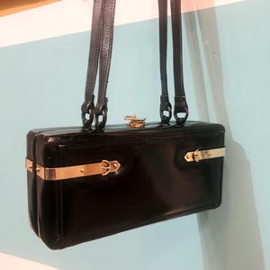 Puede incluir: Un bolso de mano rectangular negro con herrajes dorados, que incluyen un cierre y barras decorativas. El bolso tiene dos correas largas y finas y parece estar hecho de un material brillante. El fondo es una pared azul claro y blanca.