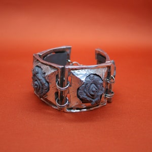 Könnte beinhalten: Ein silbernes Armband mit einem Scharnierdesign mit drei erhabenen, grauen rosenförmigen Details.