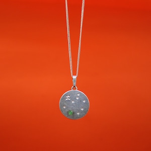 Puede incluir: Un collar con colgante plateado con un dije redondo con diseños de estrellas y geométricos, y una pequeña piedra verde. El collar cuelga sobre un fondo naranja vibrante, mostrando los detalles de la joya.