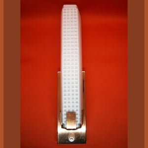 Op de afbeelding: Een witte, matglazen wandlamp met een messing frame. De lamp heeft een vierkant rasterpatroon en een enkele gloeilamp.