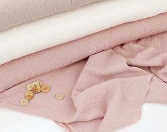 May include: A stack of three different colored fabric swatches. The top swatch is a light beige, the middle swatch is white, and the bottom swatch is a light pink. There are several wooden buttons scattered on the pink fabric.